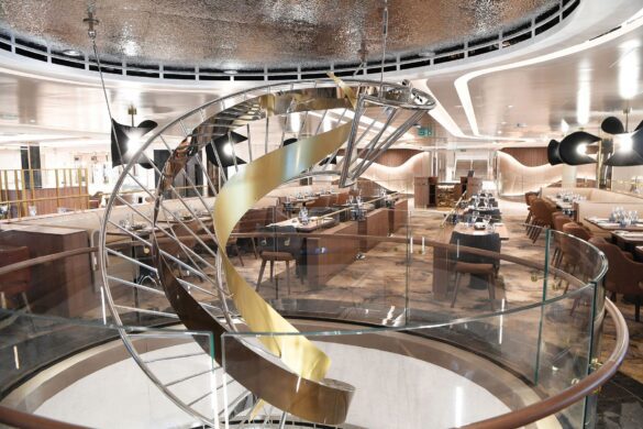 Modern restaurant interior with a spiral staircase featuring a gold ribbon-like sculpture, glass railing, and neatly arranged dining tables in the background.