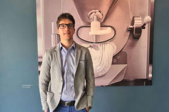 A man wearing glasses and a gray blazer stands in front of a large photo of an industrial mixing machine on a blue wall.