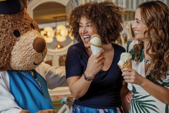 Two women smile and hold ice cream cones while standing next to a person in a bear costume inside a brightly lit room.
