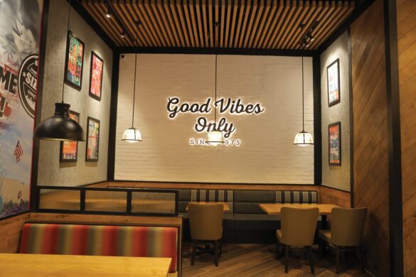 Restaurant seating area with wooden tables, cushioned booths, chairs, framed wall art, and a neon sign reading "Good Vibes Only" on a white brick wall.