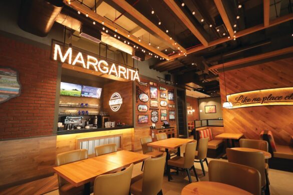 A modern restaurant interior with wooden tables and chairs, a bar area, neon signs reading "Margarita," and string lights on the ceiling.