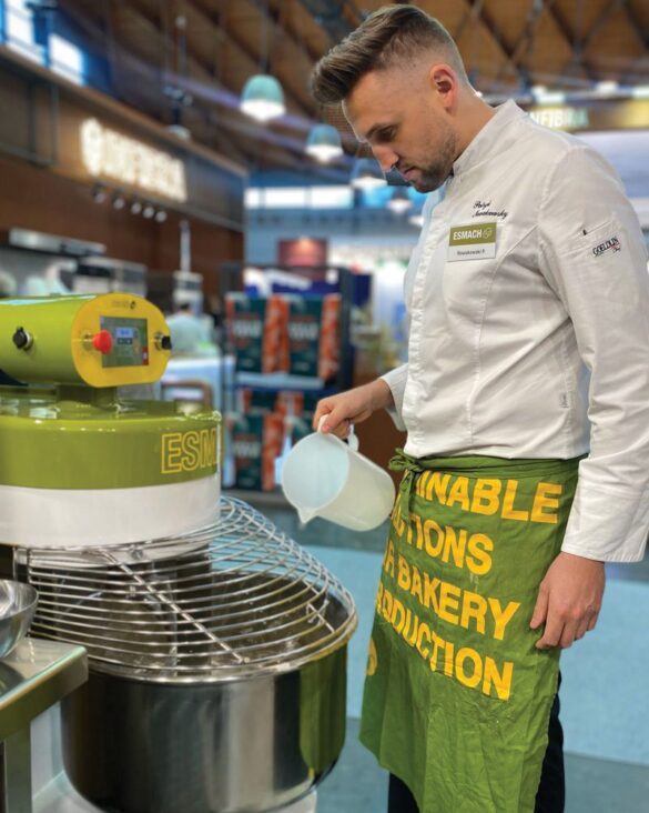 A man in a chef’s uniform pours liquid from a plastic jug into a large industrial mixer in a bakery setting.