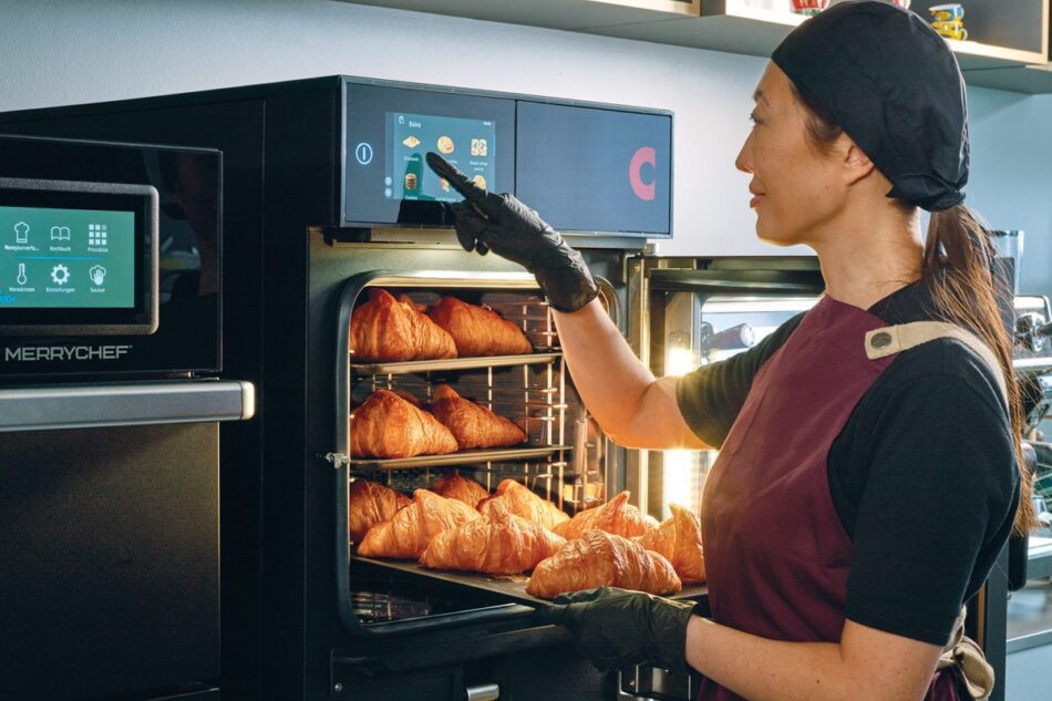 A woman wearing a uniform and gloves operates a touchscreen oven while baking croissants on trays inside a commercial kitchen.
