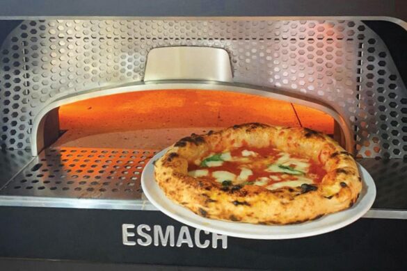 A Neapolitan-style pizza with melted cheese and basil on a white plate is placed in front of an Esmach pizza oven.