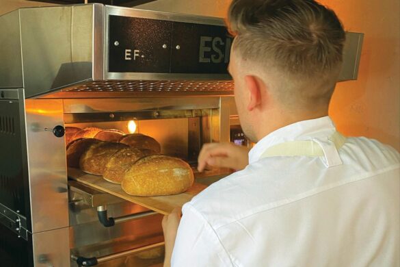 A person in a white shirt uses a wooden paddle to place several loaves of bread into a commercial oven for baking.