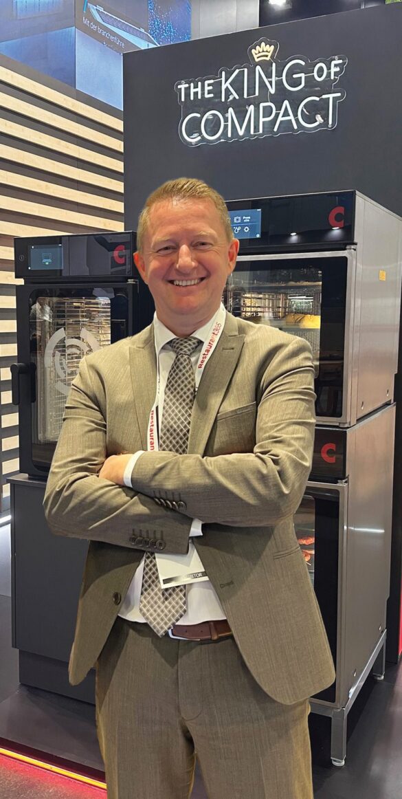 A person in a suit stands smiling with arms crossed in front of commercial kitchen appliances and a sign reading "The King of Compact.