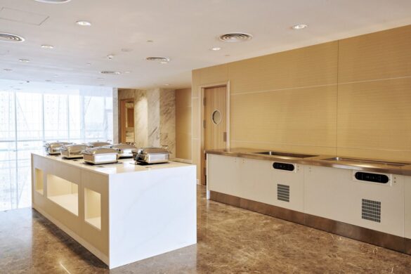 A modern, empty buffet area with chafing dishes on a central white island and stainless steel counters against beige walls.