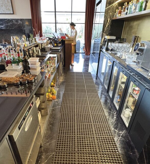 A bar with various bottles, glassware, and fruit on counters; a person stands near a window using a device.