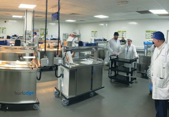A group of people in white coats and hairnets work around food service trolleys in a clean, industrial kitchen setting.