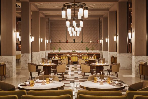 Spacious, modern restaurant interior with round tables, brown and yellow chairs, chandeliers, and decorative lighting. Marble accents and minimalist décor are visible throughout the room.