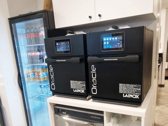 Two black Lainox Oracle commercial kitchen ovens are placed side by side on a counter, next to a glass-door fridge stocked with bottled drinks.