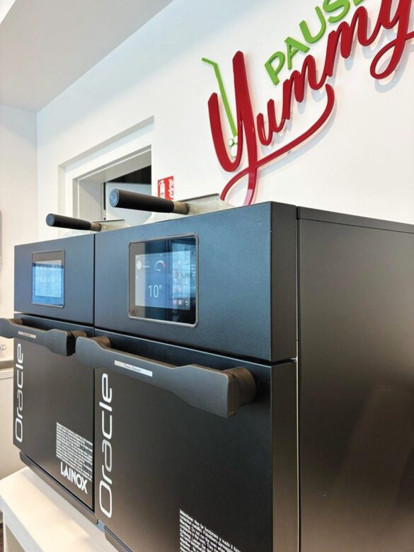 Two black Lainox Oracle ovens with digital screens are placed below a wall sign that reads "PAUSE Yummy" in green and red lettering.