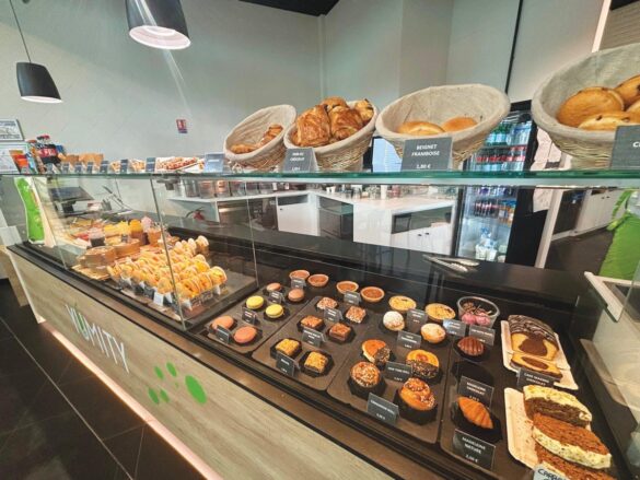 Bakery display case featuring assorted pastries, tarts, breads, and desserts in baskets and trays; refrigerated drinks visible in the background.