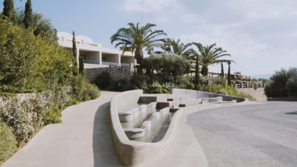 Curved concrete seating area with built-in water feature, surrounded by greenery and palm trees, near a modern building under a blue sky.