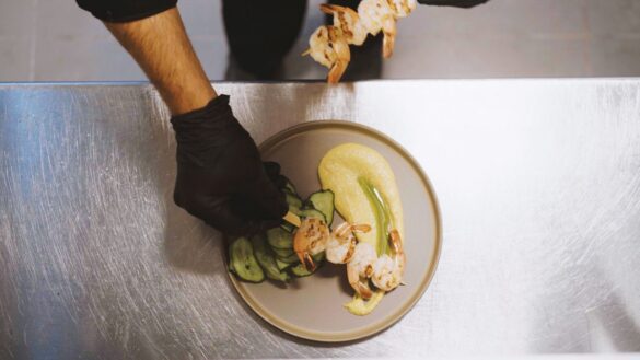 A person wearing black gloves plates grilled shrimp skewers with sliced cucumbers and a yellow puree on a round dish.