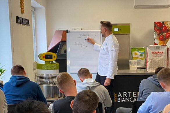 A chef in a white coat teaches a class, writing on a flip chart. Several students sit facing him in a kitchen setting with baking equipment and ingredient bags visible.