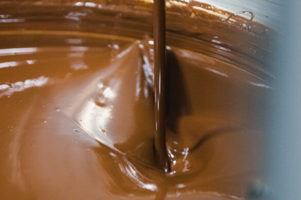 A close-up of melted chocolate being poured into a container, creating ripples and swirls on the smooth surface.