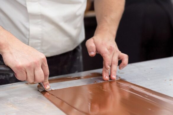 A person uses a metal scraper to spread and shape melted chocolate on a metal surface.