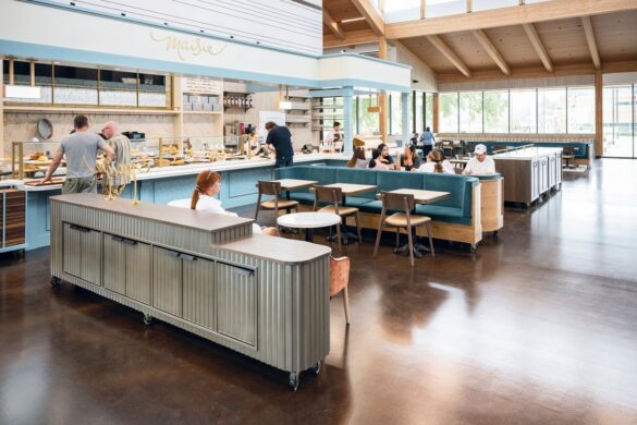 Modern, spacious cafe with teal booths and open kitchen; several people are seated, with staff preparing food behind the counter under natural light from large windows.