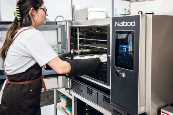 A person wearing gloves places a tray of food into a Naboo commercial oven in a professional kitchen setting.