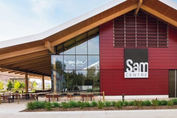 Exterior view of the Sam Centre building with red siding, large windows, and visible signage for “Sam Centre” and “Yardie Eatery.” Outdoor seating and landscaping are also visible.
