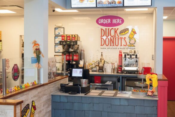 Duck Donuts counter with coffee machines, syrup bottles, a register, and “Order Here” sign on the wall, with autumn decorations and a tray of small rubber ducks on the counter.