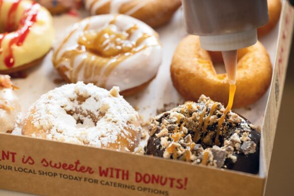 A hand drizzles caramel sauce onto a chocolate donut topped with crumbs, surrounded by assorted donuts in a box.
