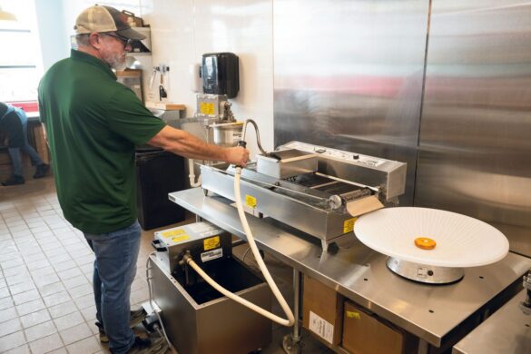 A man operates a donut-making machine in a commercial kitchen, with one finished donut on a white tray beside the equipment.