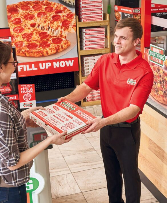 A store employee in a red shirt hands a pizza box to a customer inside a pizza shop with pizza boxes and posters in the background.