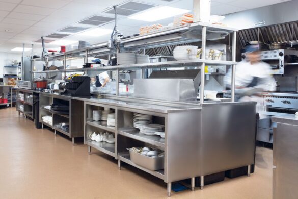 A stainless steel commercial kitchen with shelves, stacked plates, utensils, and two blurred figures in chef uniforms working.
