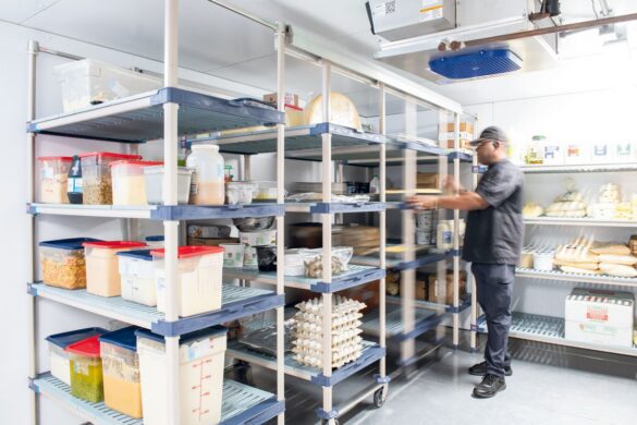 A person organizes shelves filled with food containers, eggs, and bread inside a commercial walk-in refrigerator.