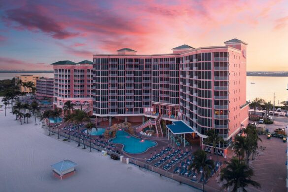 A large pink beachfront resort with multiple floors, an outdoor pool, rows of lounge chairs, palm trees, and a sunset sky in the background.